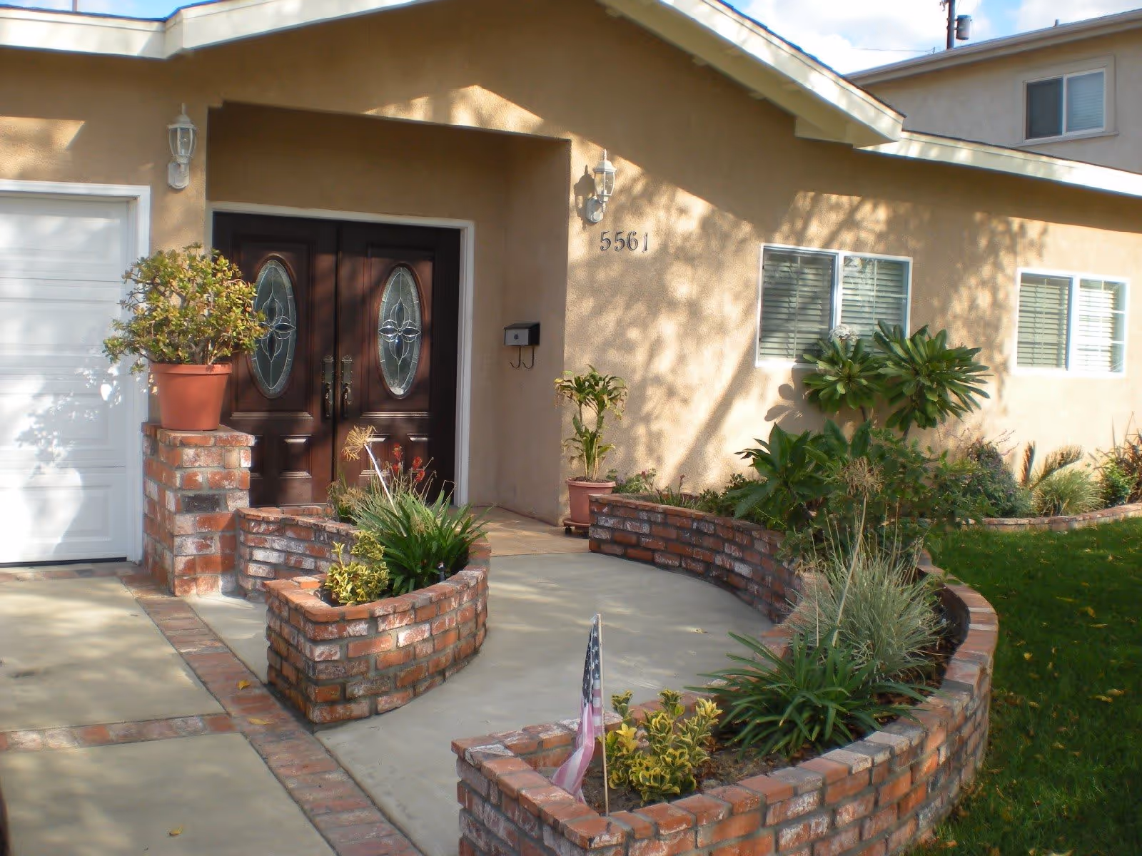 Front entrance of a single-story house with double decorative wooden doors, brick planter walls, and a small landscaped yard.