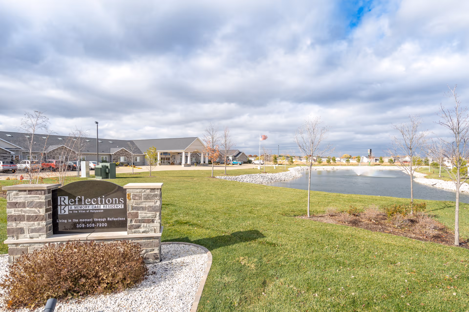 Exterior view of Reflections Memory Care residence in Washington, IL, showing a stone sign with the facility name and phone number, a large green lawn, a pond with a fountain, several young trees, and a building with a covered entrance under a cloudy sky.