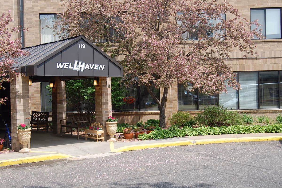 Front entrance of the Wellhaven Senior Living building with a covered canopy, benches, potted plants and a flowering tree.