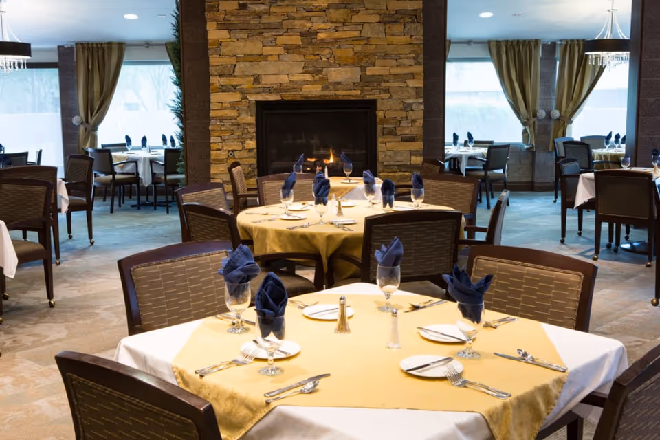 A dining room with multiple tables set with white and yellow tablecloths, blue folded napkins in glasses, and silverware. The room features a stone fireplace in the center with a fire burning, large windows with green curtains, and several chairs around each table.