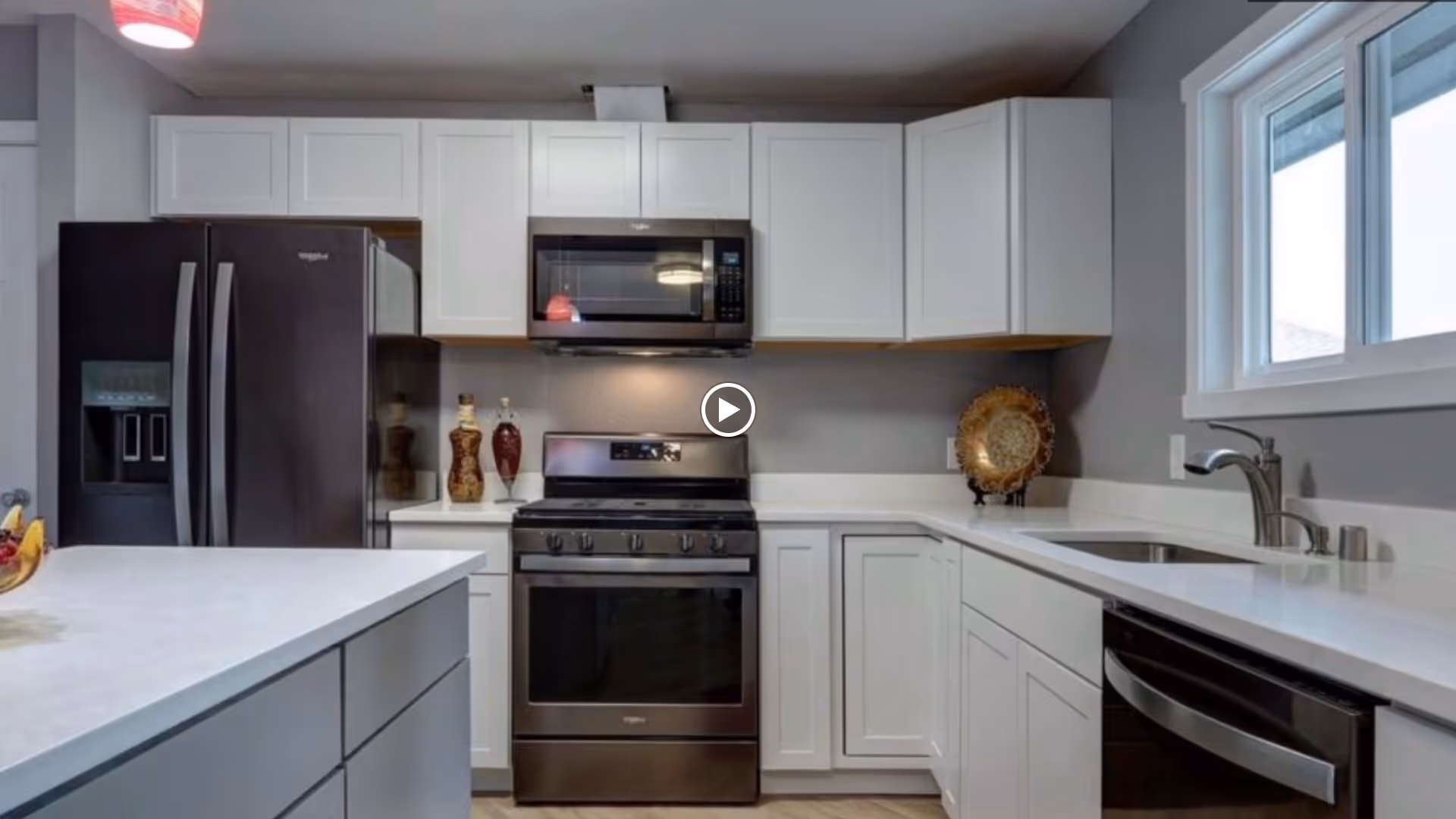 Modern kitchen with white cabinets, a black refrigerator, a black microwave above a black stove, a white countertop with a sink, and a window letting in natural light.