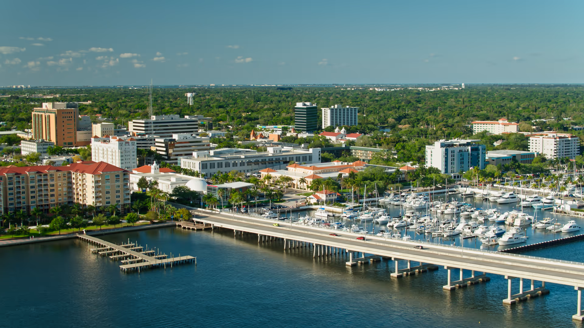Aerial view of a coastal city with a marina, bridge, and green skyline.