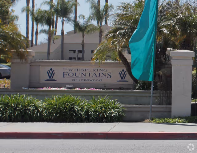 Entrance sign for Whispering Fountains at Lakewood with palm trees and landscaping.