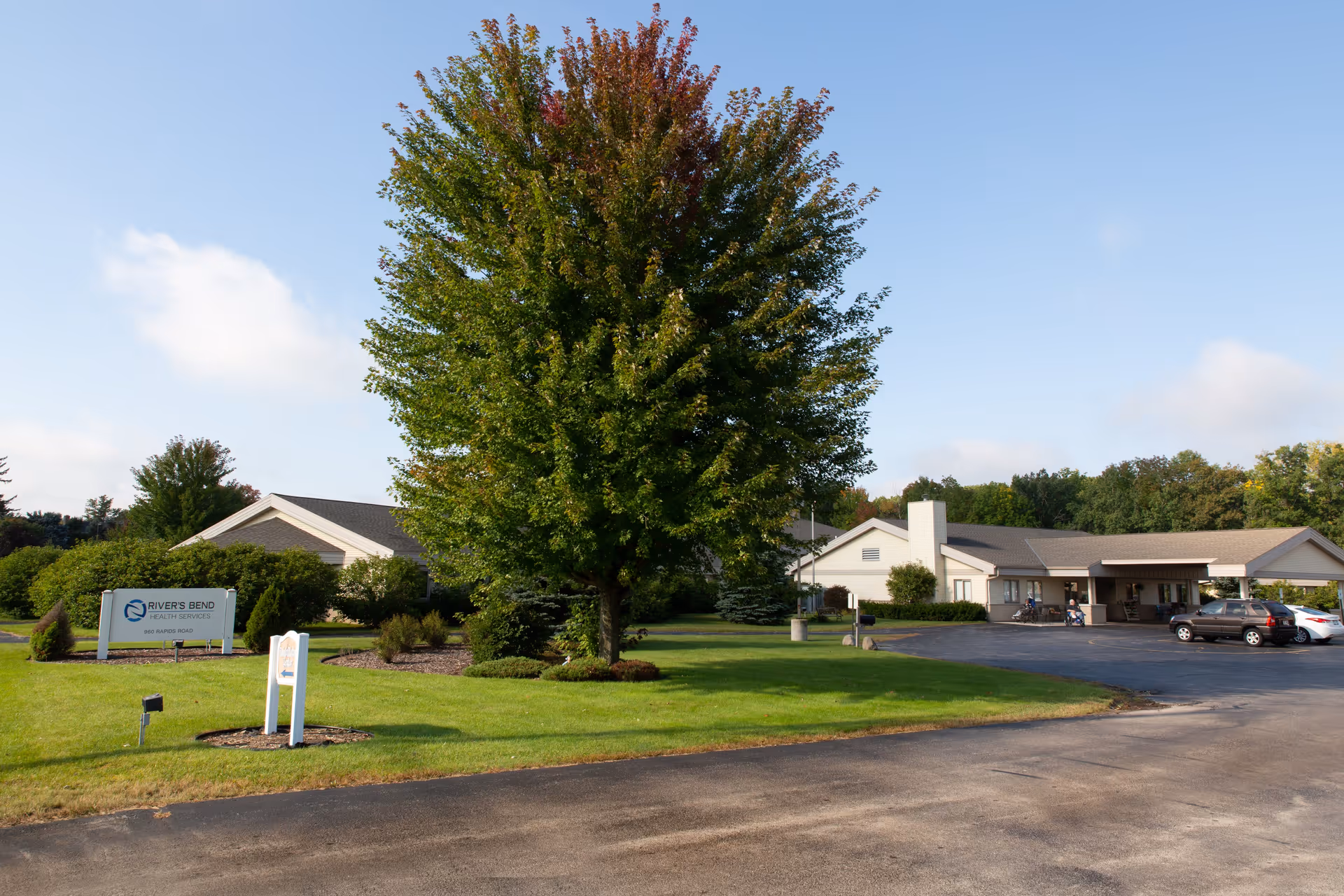 Front exterior of River's Bend Health Services single-story building with a large tree and manicured lawn in the foreground and a parking area to the right.