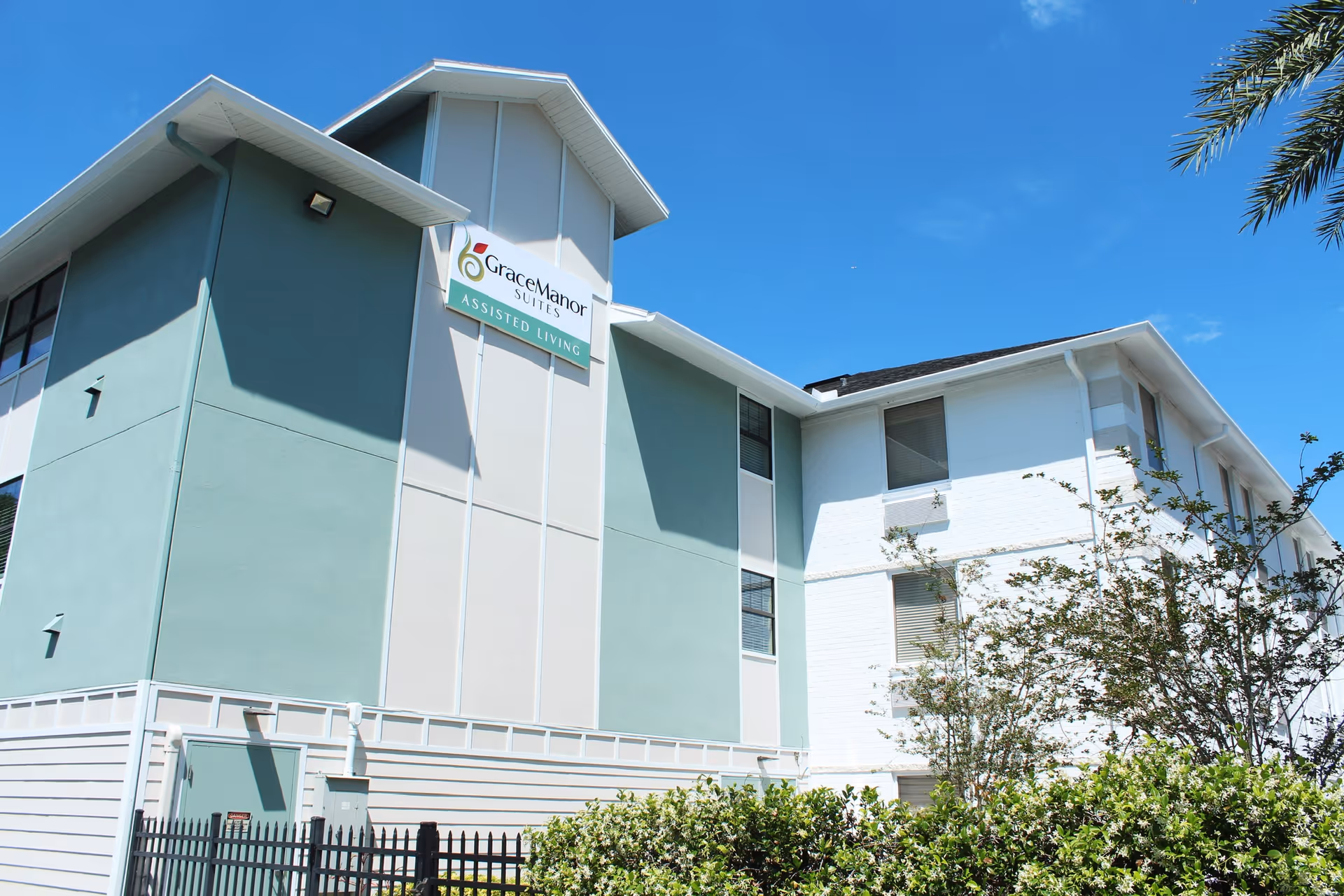 Exterior view of a multi-story assisted living facility named Grace Manor Suites under a clear blue sky, with green and white walls, windows, and surrounding bushes and trees.