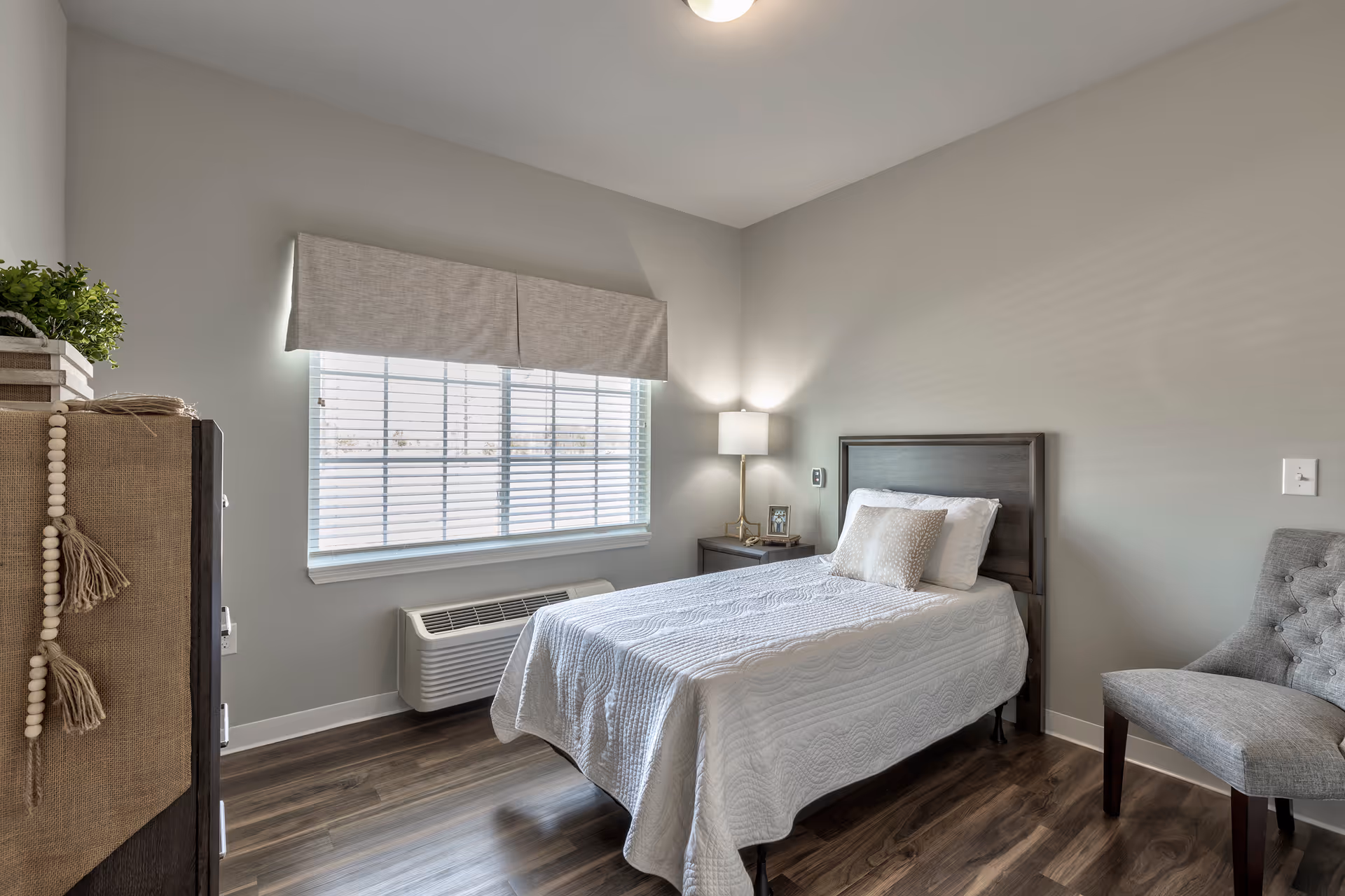 A clean bedroom in a senior living facility with a bed, nightstand, chair, and window.