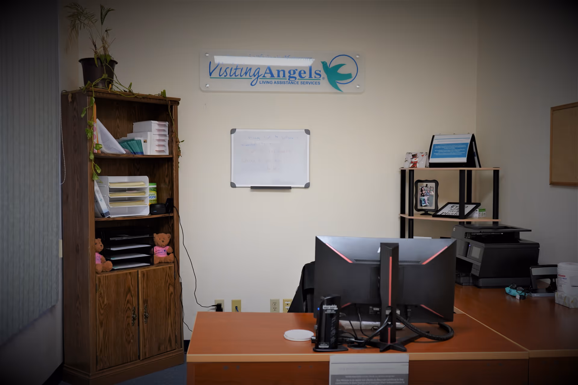 Office reception area with desk, computer, shelves, and a Visiting Angels sign.