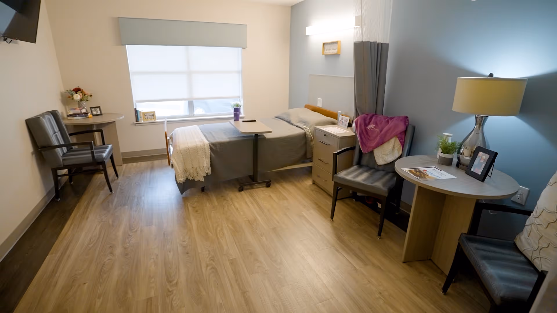 A clean and well-lit senior living bedroom with a single bed covered in gray bedding and a white knitted throw. The room features wood flooring, two black chairs, a small round table with a lamp, a plant, and framed photos. A window with a white shade lets in natural light, and a TV is mounted on the wall.