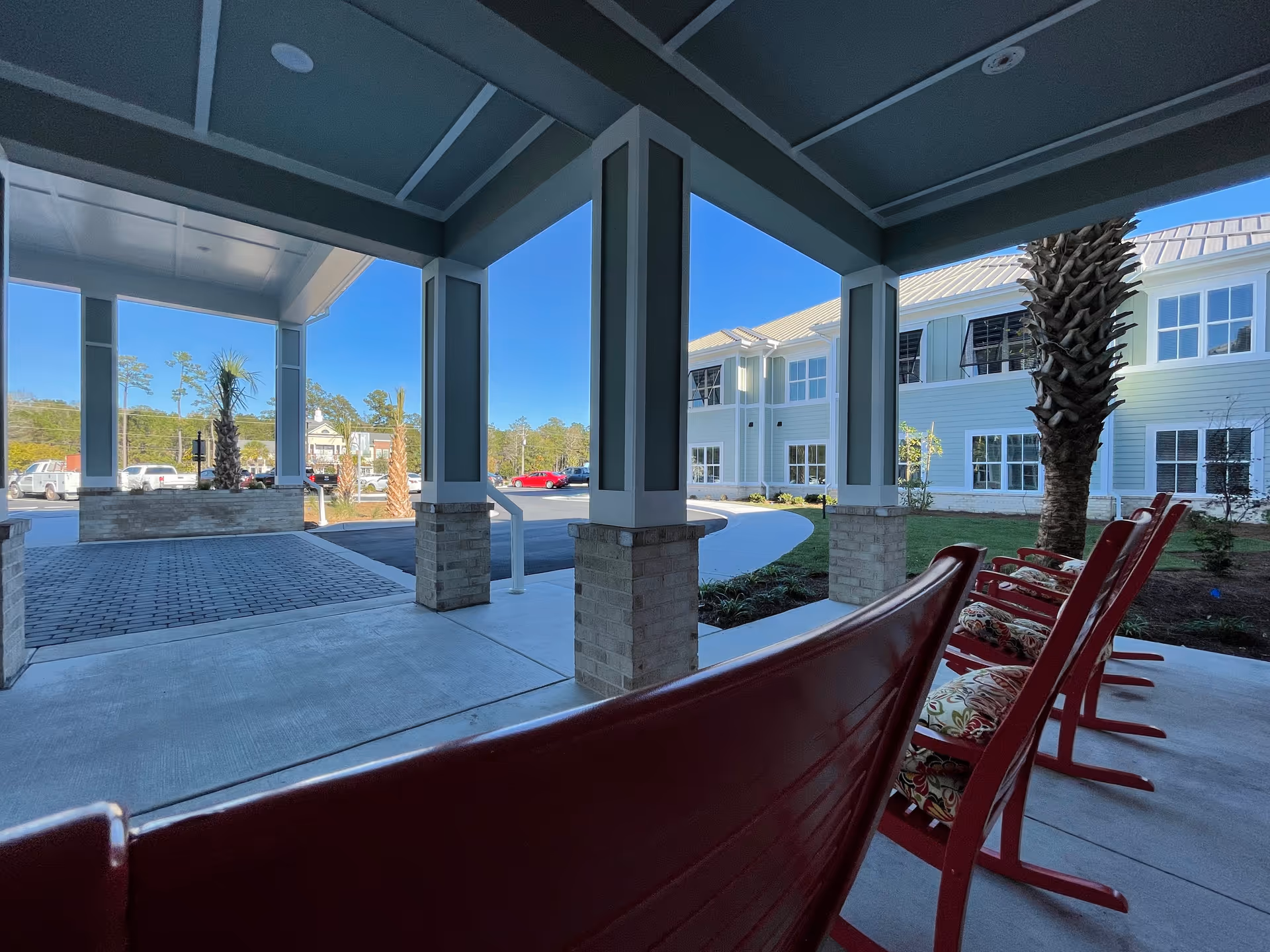 Covered outdoor seating area with red rocking chairs and cushions, overlooking a driveway and a two-story building with light blue siding and white trim under a clear blue sky.