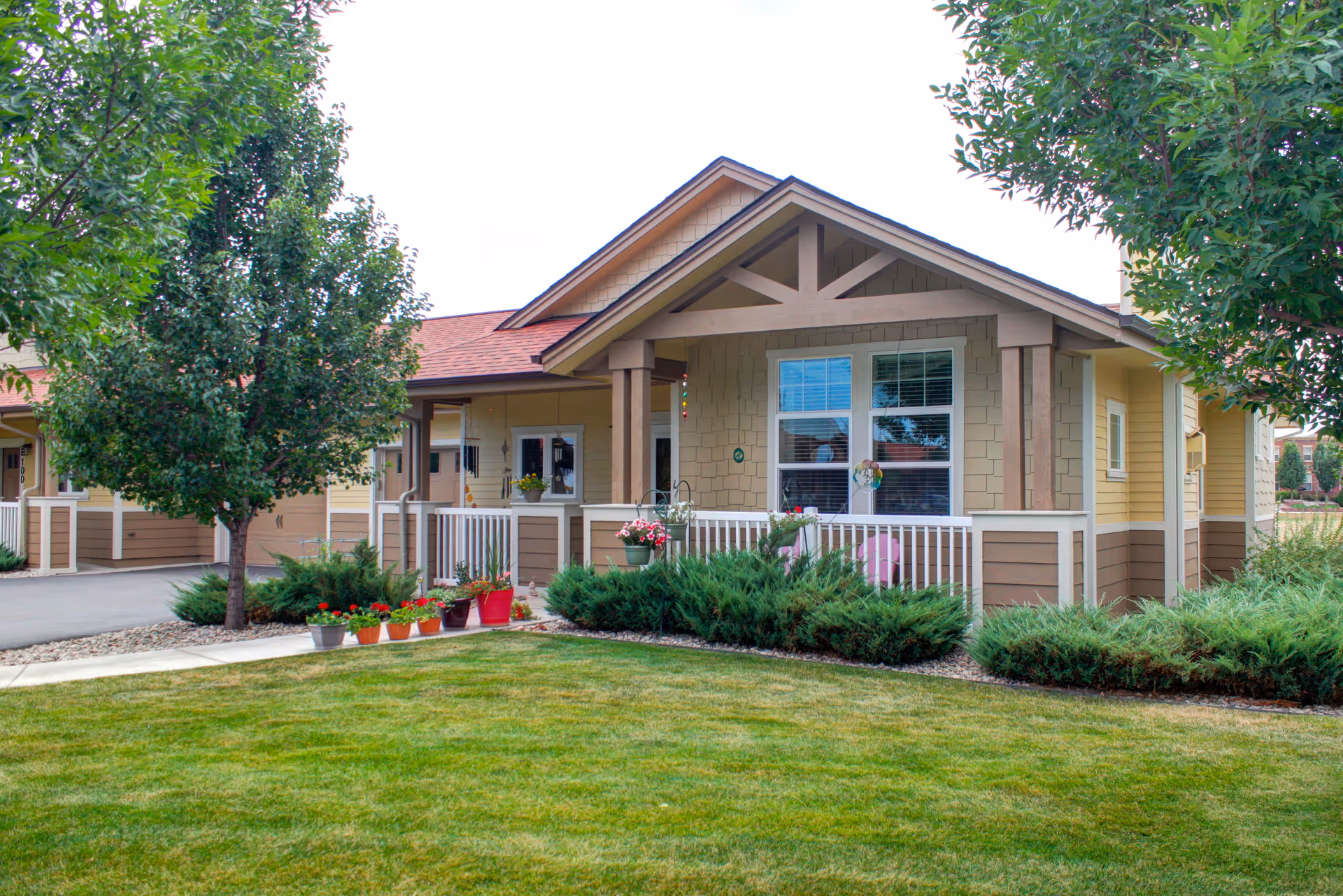 Exterior view of a single-story senior living facility building with a covered porch, white railing, and several potted plants. The building has beige and brown siding with a peaked roof. There are green bushes and a well-maintained lawn in the foreground, along with two trees on either side.