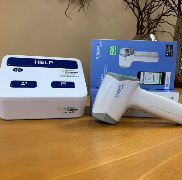 A close-up of a HELP electronic caregiver alert device and thermometer on a wooden table.