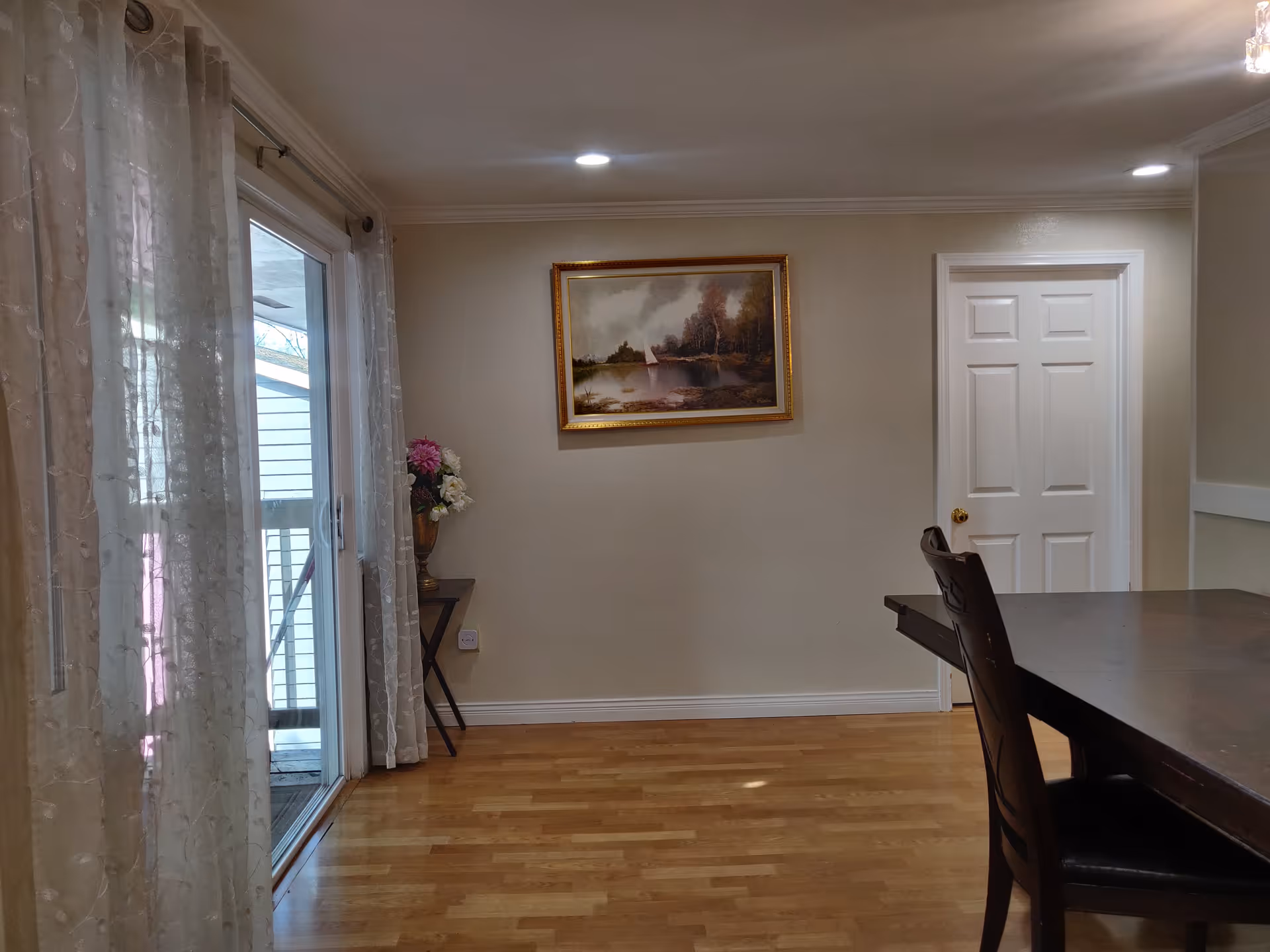 Interior view of a room with wooden flooring, a dark wooden dining table with chairs on the right, a white door on the back wall, a framed painting of a landscape above a small table with a vase of flowers, and sheer curtains covering a sliding glass door on the left.