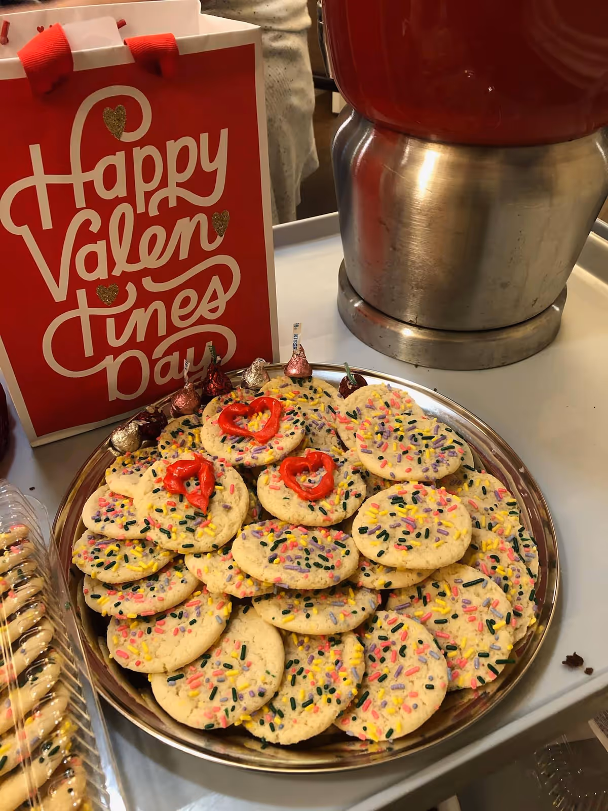 A plate filled with sugar cookies topped with colorful sprinkles and a few decorated with red heart-shaped icing. Behind the plate is a red and white gift bag with the words 'Happy Valentines Day' written on it. Part of a large metallic container is visible on the right side of the image.