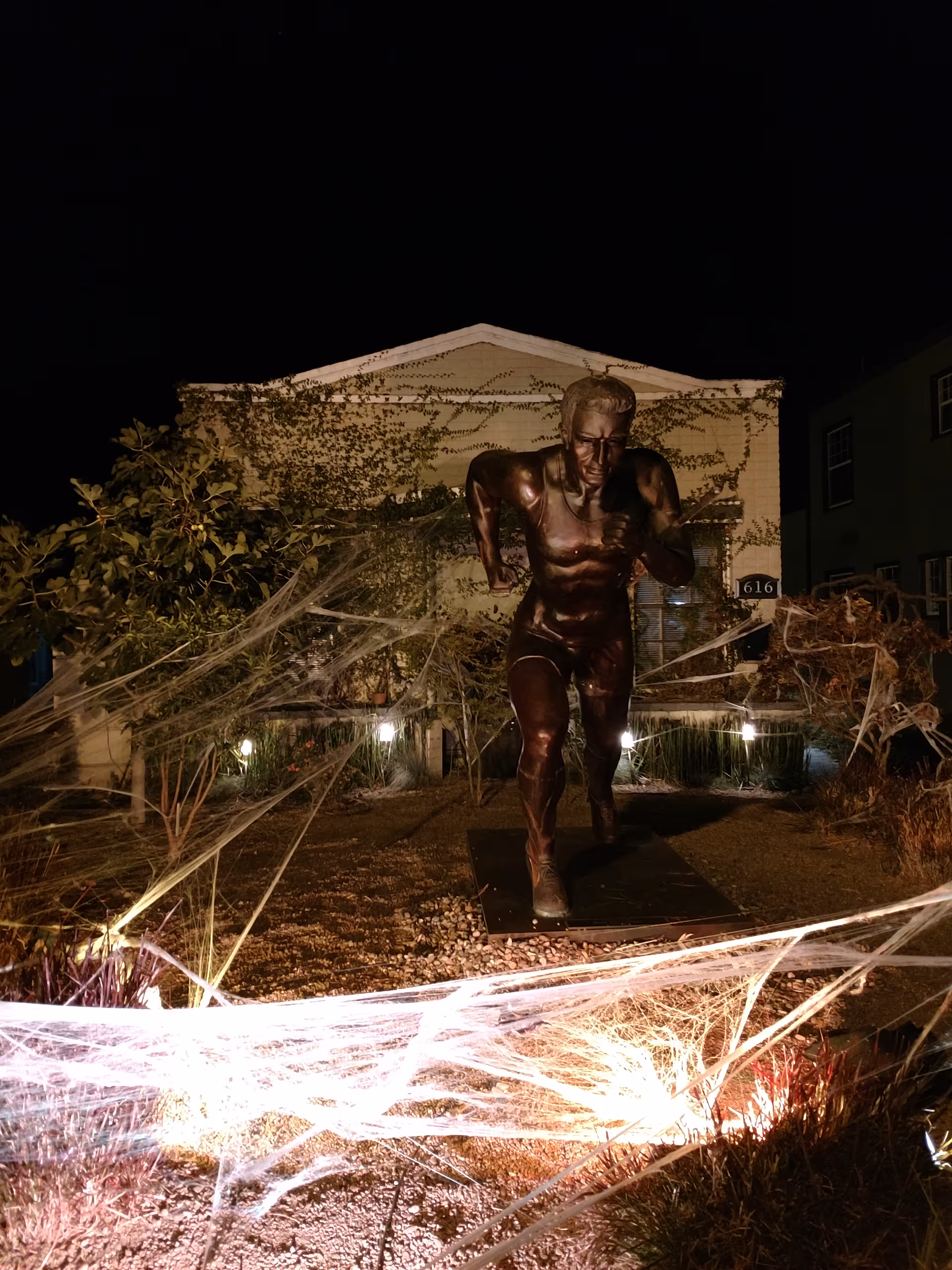 Night exterior of Webster House with a bronze athlete statue in front and spider web lights.