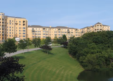Exterior view of a cream-colored senior living building with a green lawn and pond.