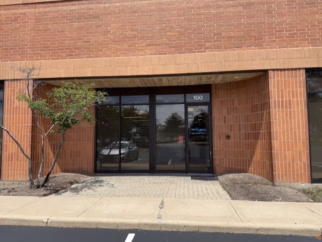 Brick storefront entrance with glass doors labeled 100, sidewalk and a small tree.