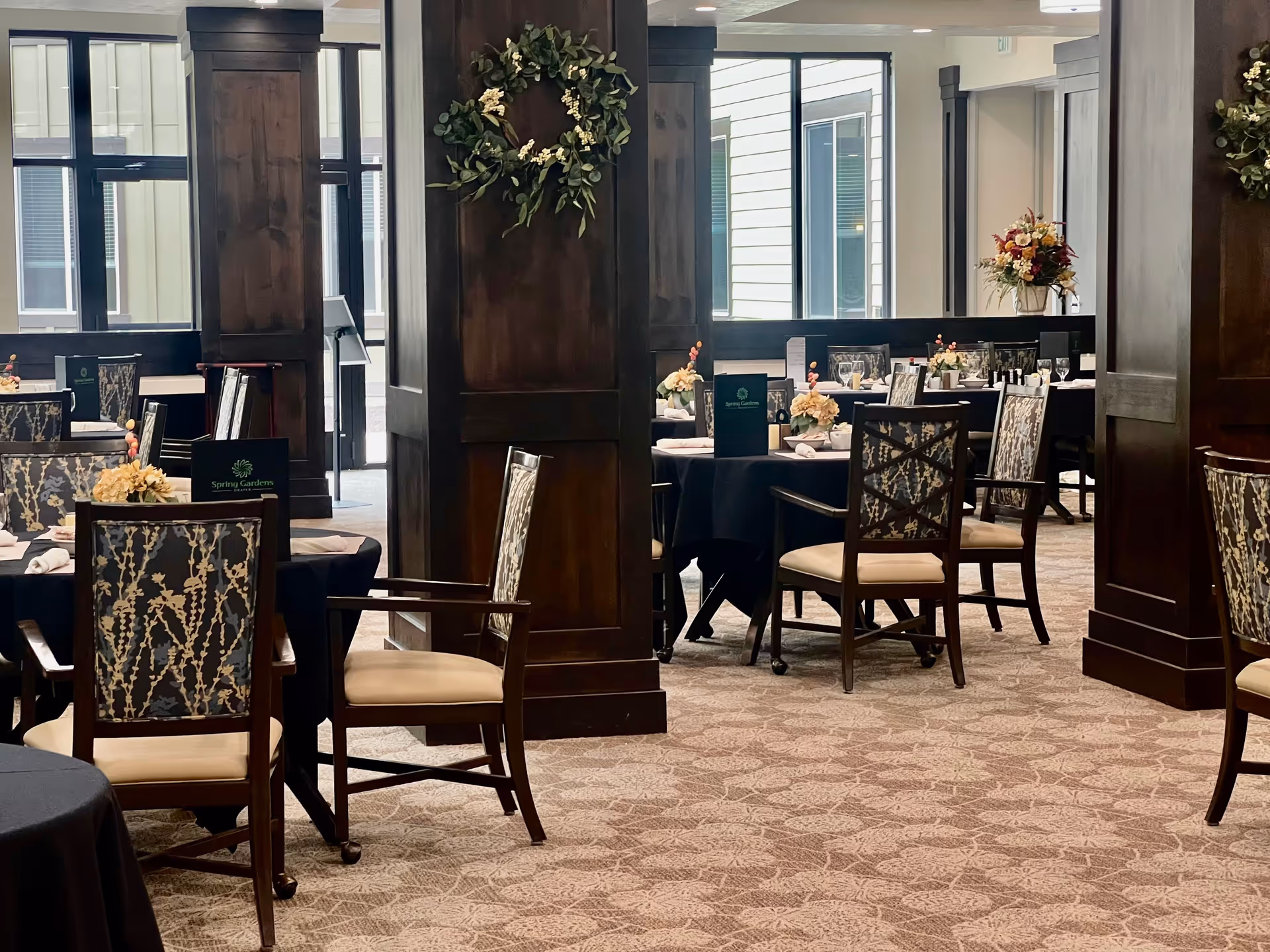 Elegant dining room with round tables covered in black tablecloths, each set with floral centerpieces, menus, and glassware. The room features dark wooden pillars decorated with green wreaths, patterned carpet, and large windows letting in natural light.