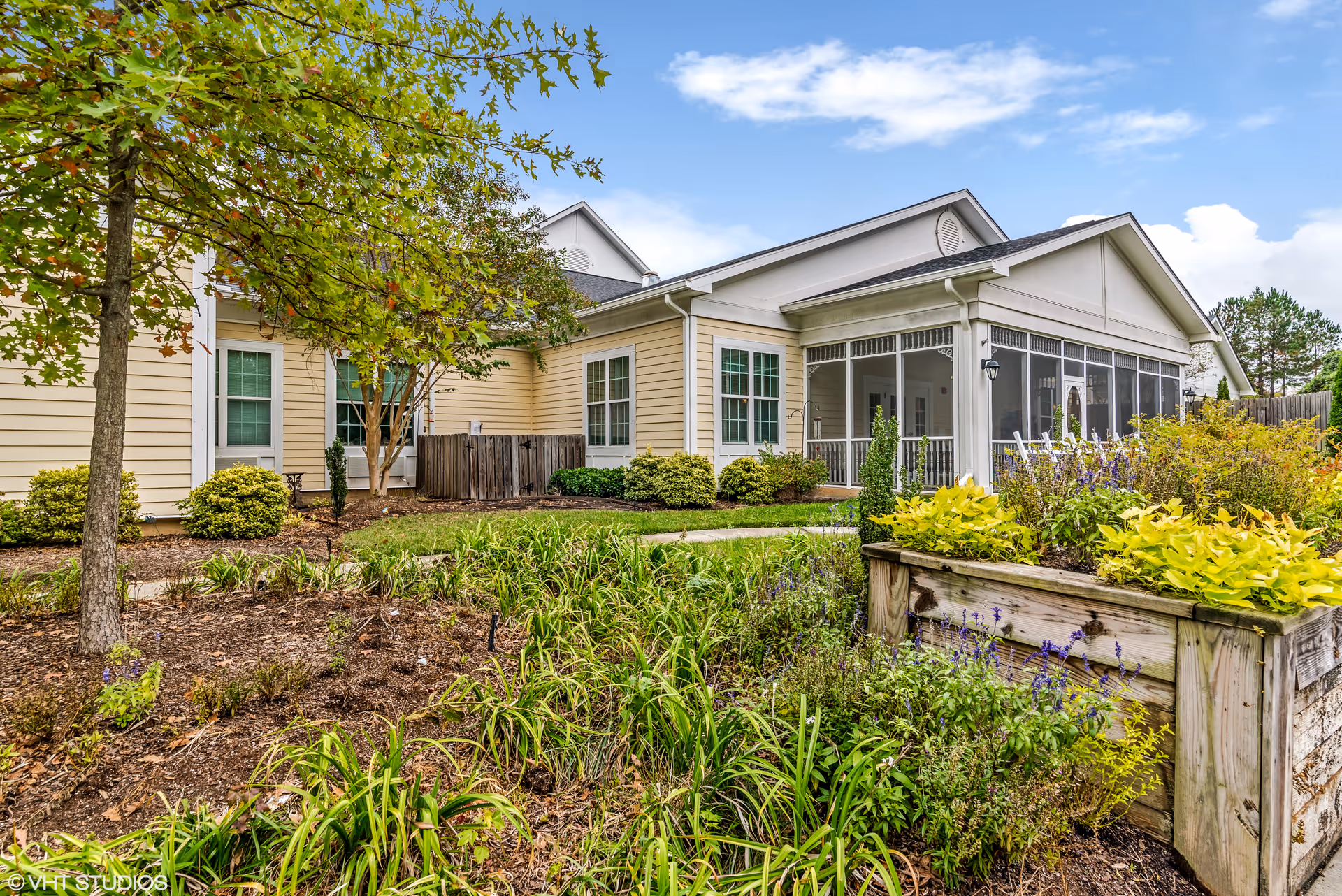Exterior view of a single-story building with beige siding and white trim, surrounded by landscaped garden beds with various plants and shrubs. A screened porch with white rocking chairs is visible on the right side of the building under a clear blue sky with some clouds.