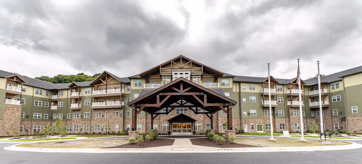 Front exterior view of a large, multi-story senior living facility named Harmony at Morgantown, featuring a covered entrance with wooden beams, multiple balconies, and a combination of stone and green siding under a cloudy sky.