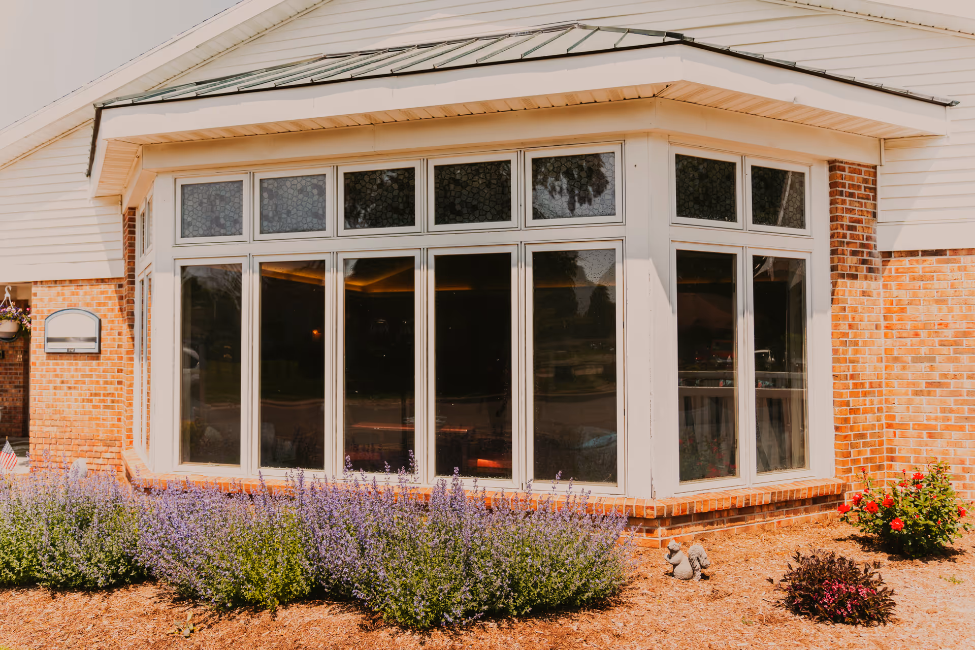 Exterior view of a senior living facility building with a large bay window, brick walls, and white siding. There are purple flowering plants and other greenery in front of the building.