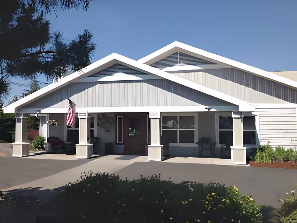 Front exterior view of Brookside Place, a single-story building with a peaked roof, white siding, and a covered entrance supported by columns. An American flag is displayed near the entrance, and there are bushes and greenery in the foreground under a clear blue sky.