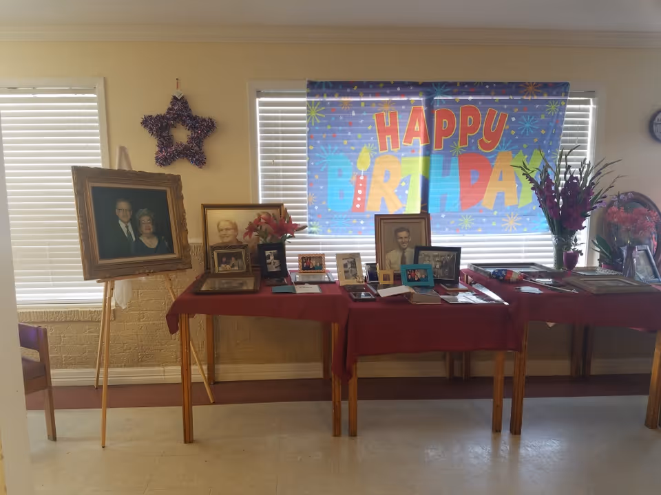 An interior room with tables displaying framed photos, flowers and decorations beneath a large 'Happy Birthday' banner.