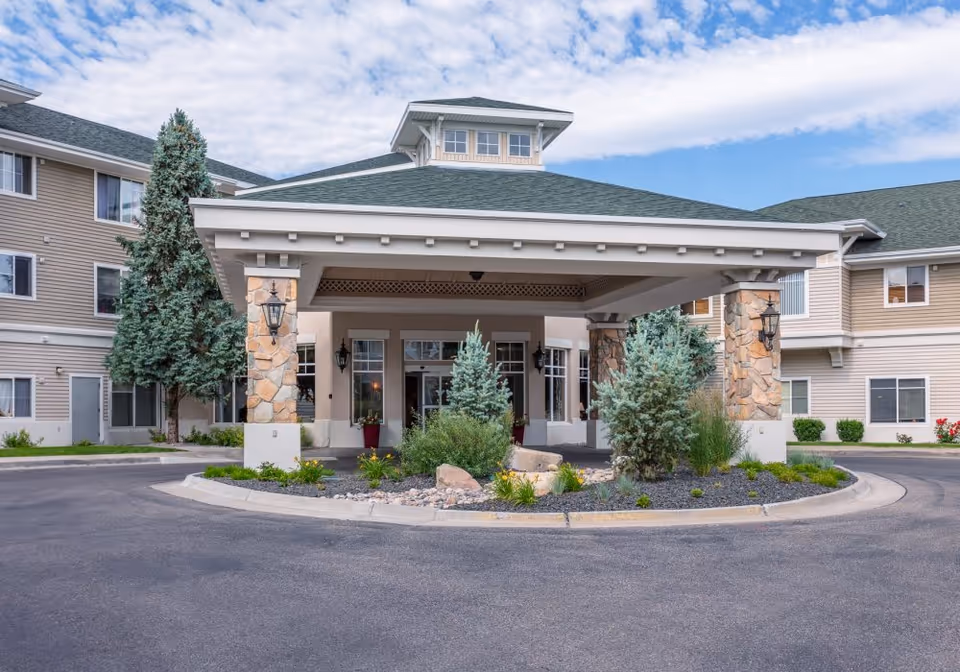 Entrance of a senior living facility with a covered driveway supported by stone pillars, surrounded by small evergreen trees and landscaping, with a multi-story beige building in the background under a partly cloudy sky.