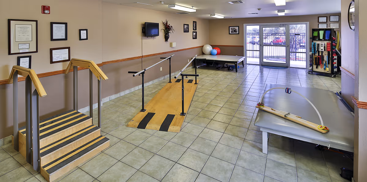 A rehabilitation therapy room with a small wooden staircase with handrails, parallel bars for walking exercises, therapy tables with exercise equipment, and exercise balls. The room has tiled floors, beige walls with a wooden chair rail, framed certificates on the wall, a wall-mounted TV, and a glass door leading outside.