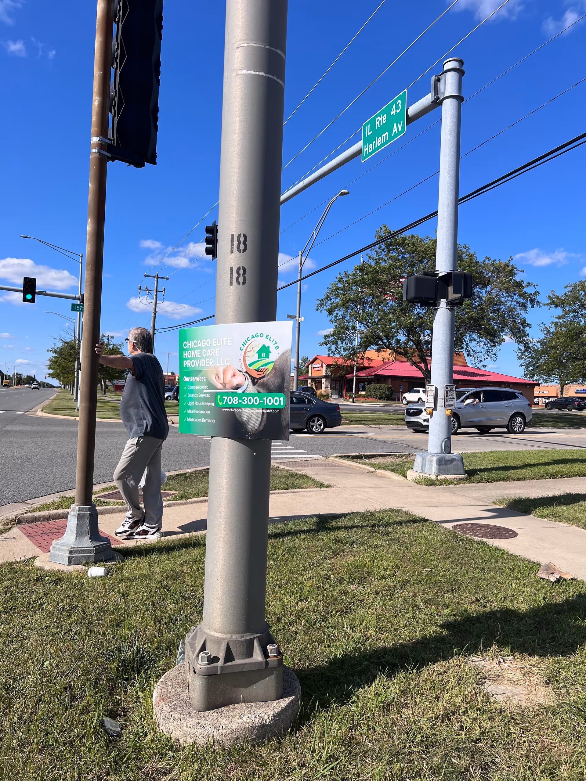 Person stands near a streetlight with a Chicago Elite Home Care poster in a sunny intersection.