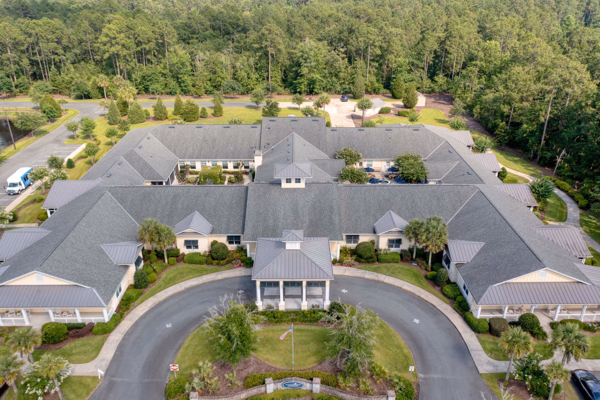 Aerial view of Addington Place of Brunswick, a large senior living facility surrounded by trees and greenery. The building has a gray roof with multiple sections and a covered entrance with a circular driveway. There are landscaped lawns, bushes, and palm trees around the building, with parking areas visible in the background.