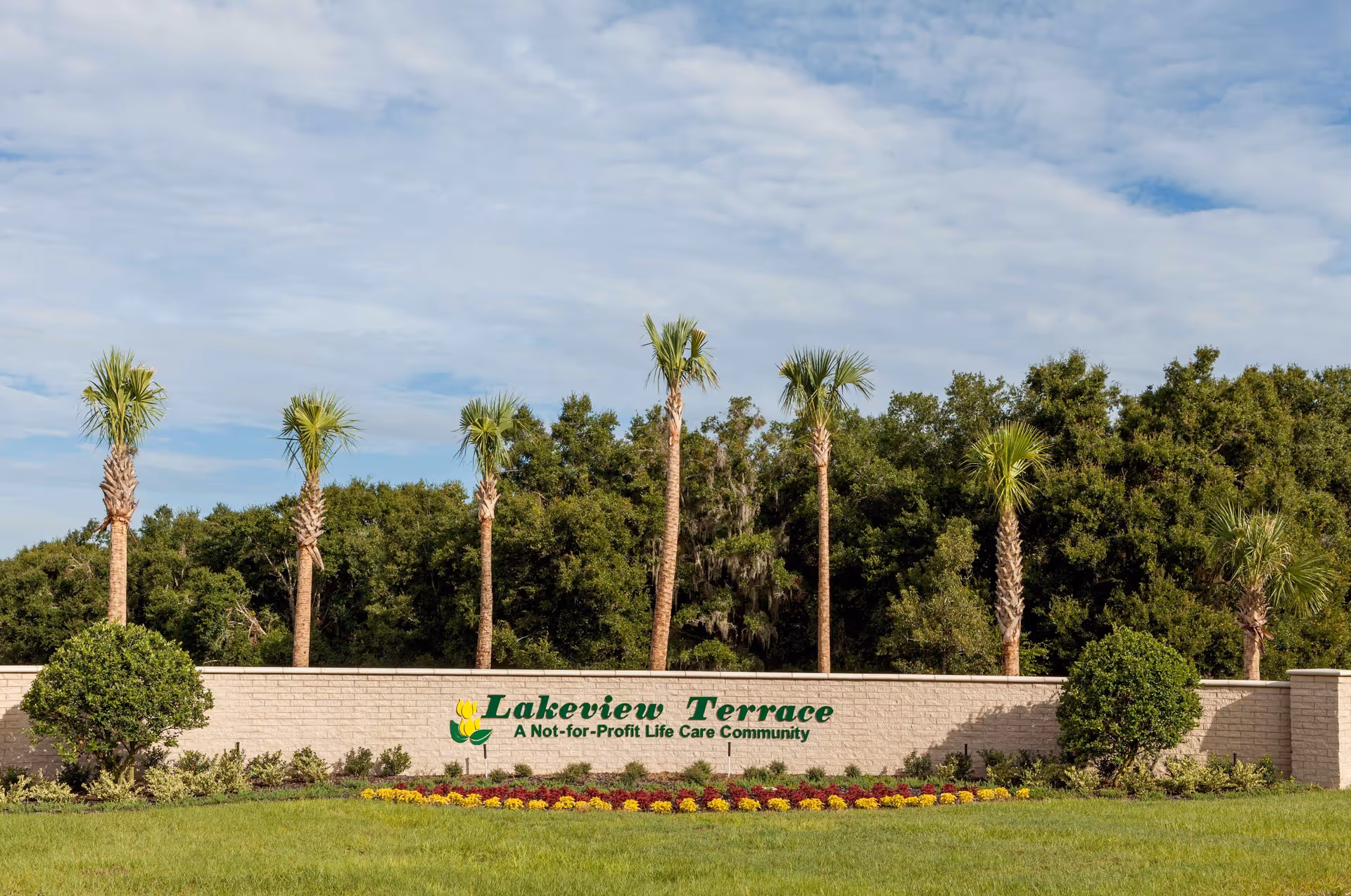 A landscaped entrance sign for Lakeview Terrace, a not-for-profit life care community, with palm trees and bushes behind a low brick wall and a flower bed in front under a partly cloudy sky.