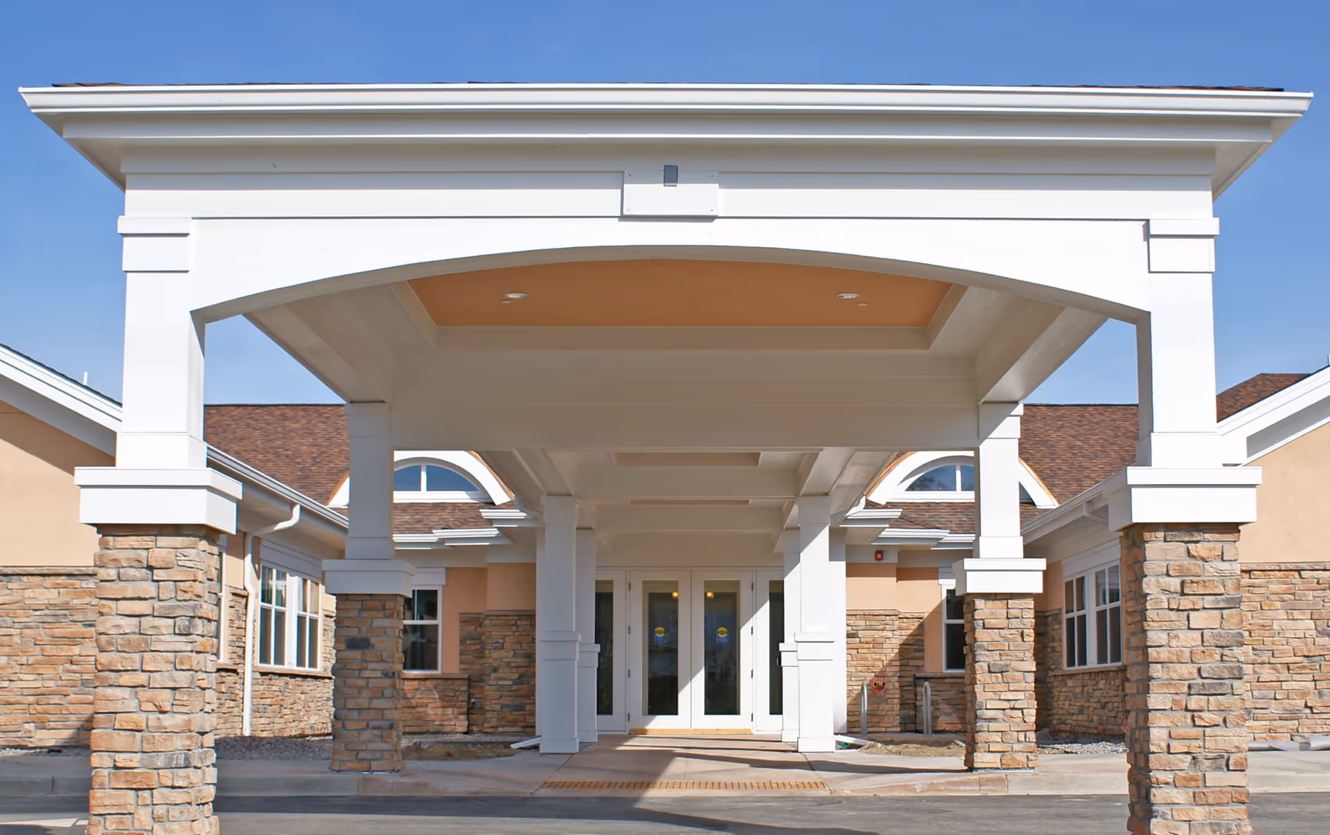 Entrance of a building with a covered drop-off area supported by white columns with stone bases, beige walls, and a brown shingled roof under a clear blue sky.