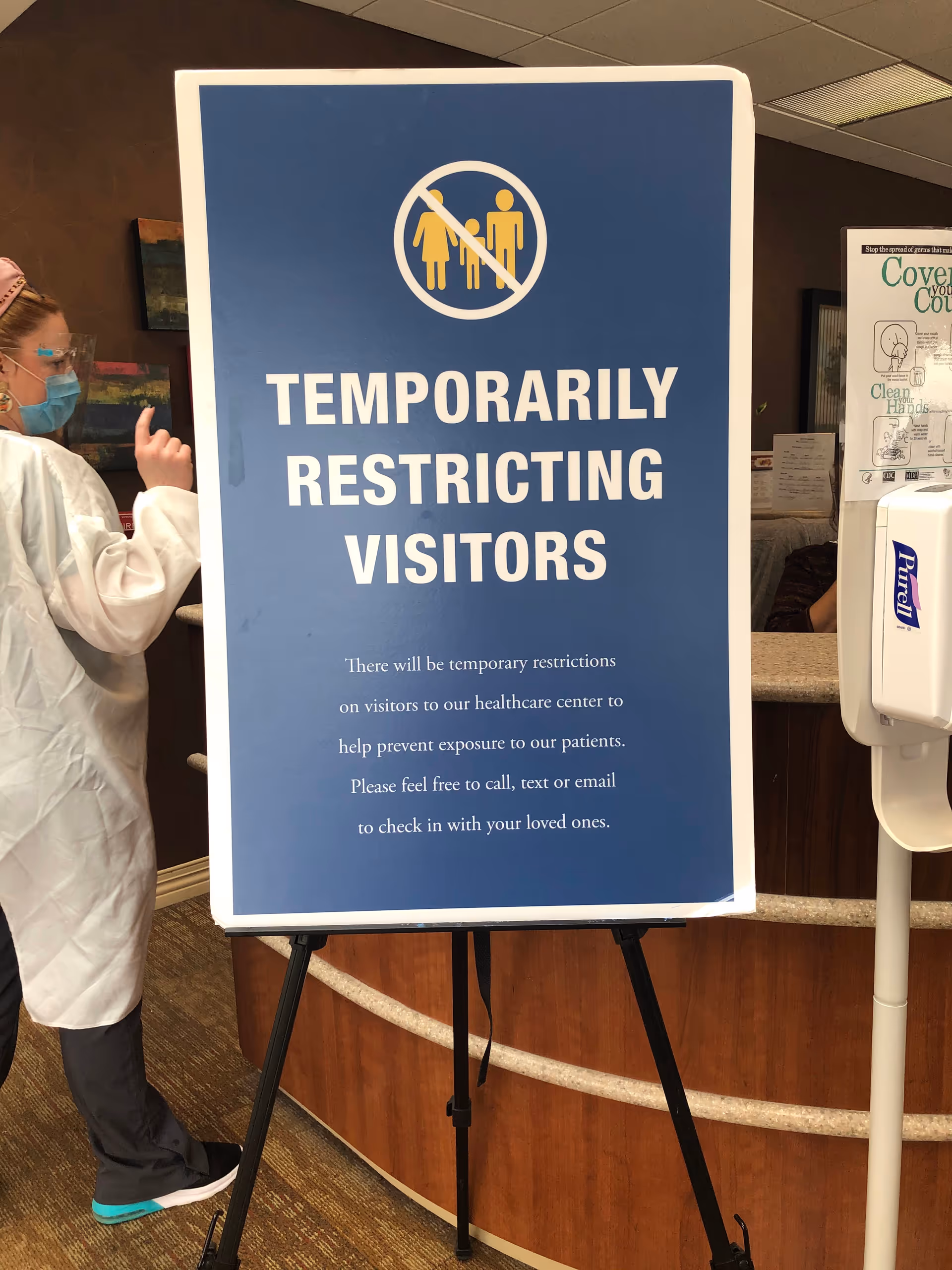 Blue sign reading Temporarily Restricting Visitors inside a healthcare facility, with a clinician nearby.