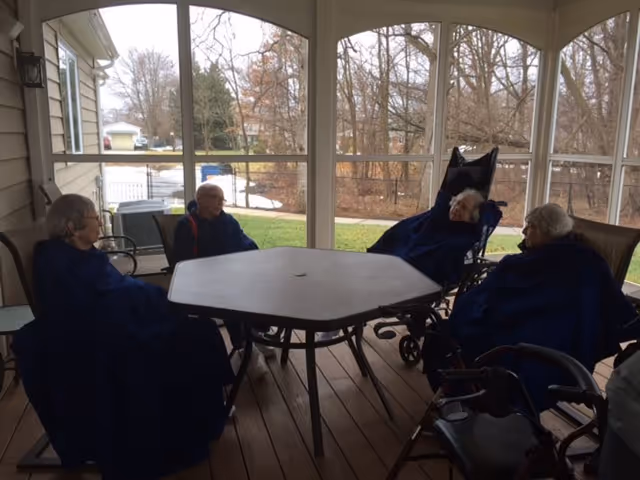 Four elderly residents sit with blankets around a patio table on a screened porch.