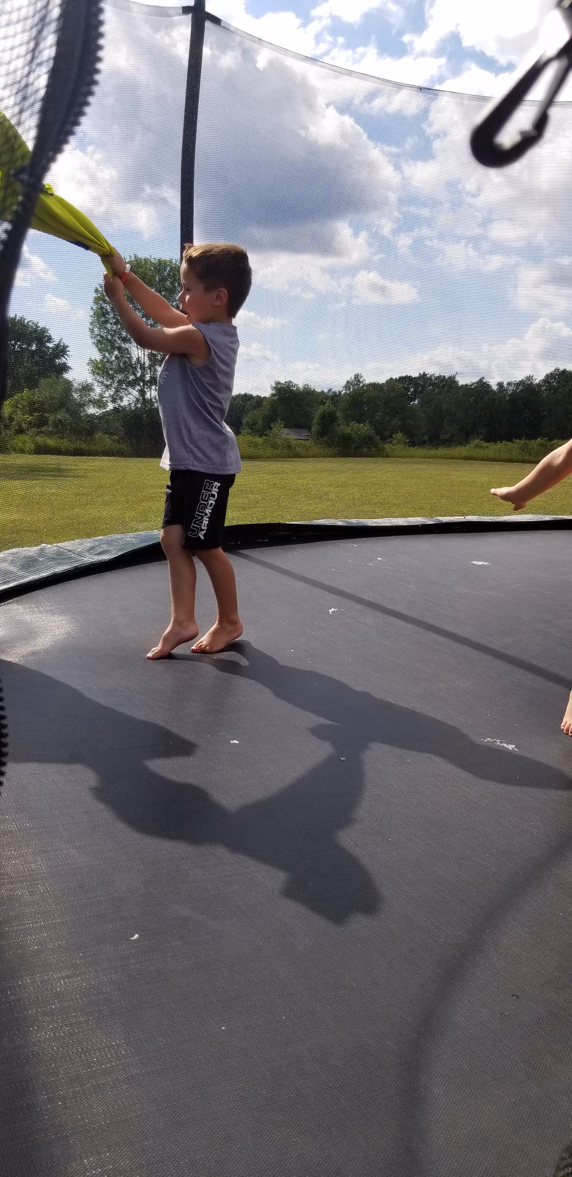 Child stands barefoot on a trampoline outdoors, grabbing a green handle with a grassy field behind.