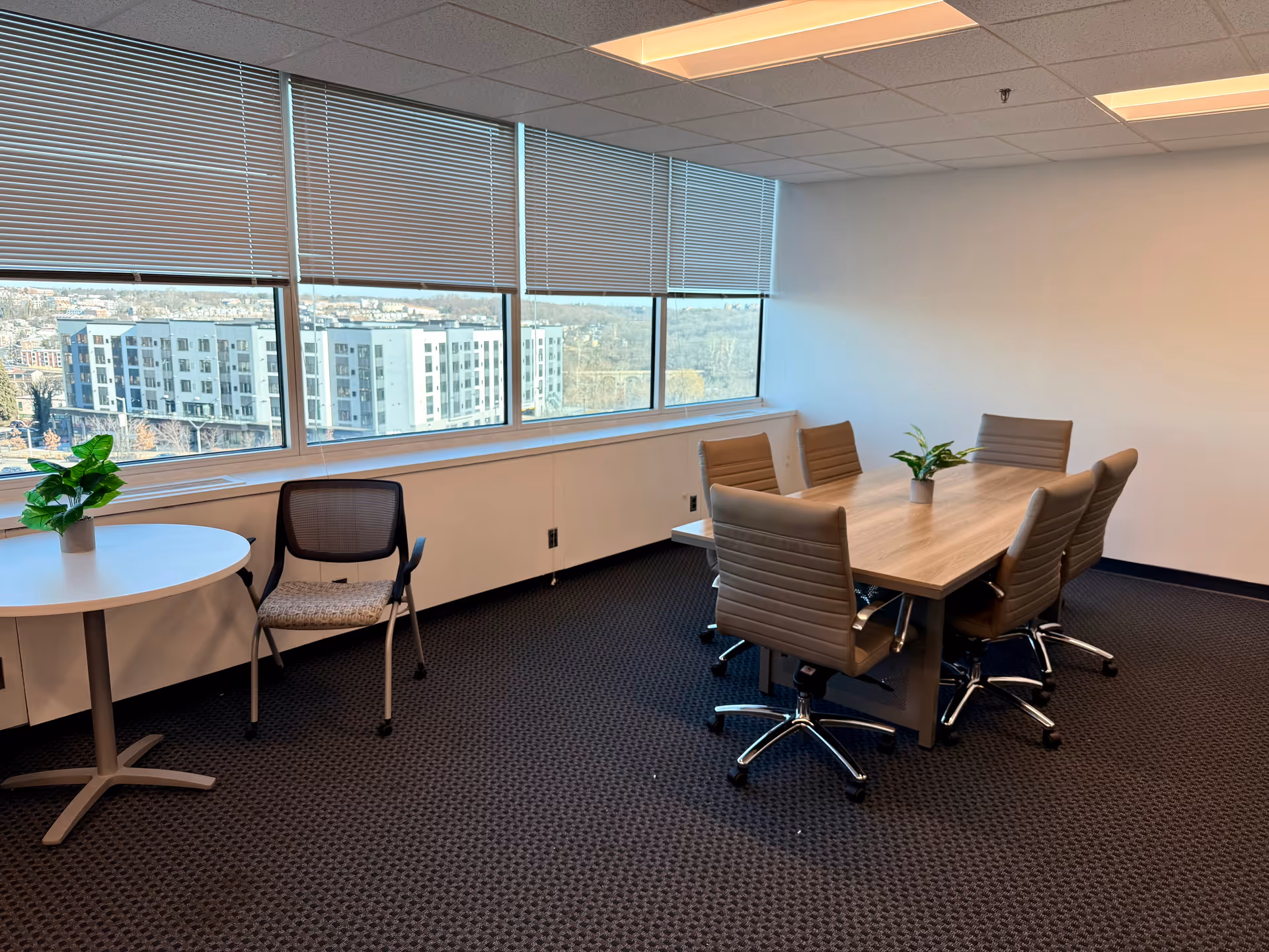 A modern interior meeting room with windows, blinds, a long table, and potted plants.
