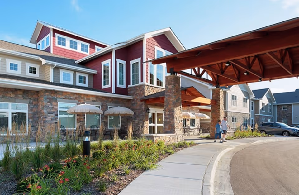 Front exterior of a senior living building with a covered stone-column entrance, outdoor seating, landscaping, and two people walking near the driveway.