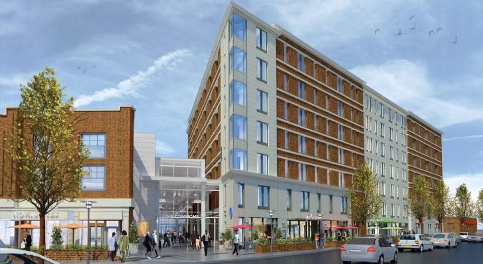 Exterior view of a modern multi-story building with brick and light-colored facade, featuring large windows and outdoor seating areas with umbrellas. People are walking and cars are parked along the street under a partly cloudy sky.