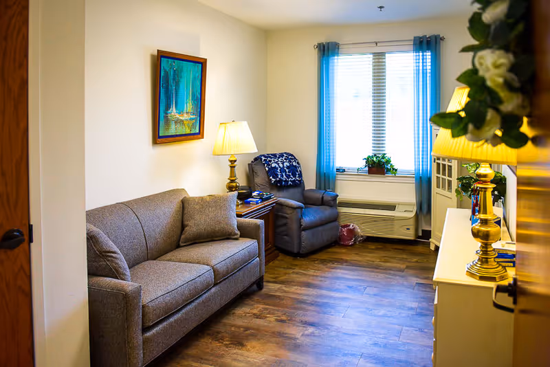 A cozy living room with a gray sofa and matching armchair, a wooden side table with a lamp, a window with blue curtains, a white cabinet with a lamp and decorative items, and a painting of sailboats on the wall.