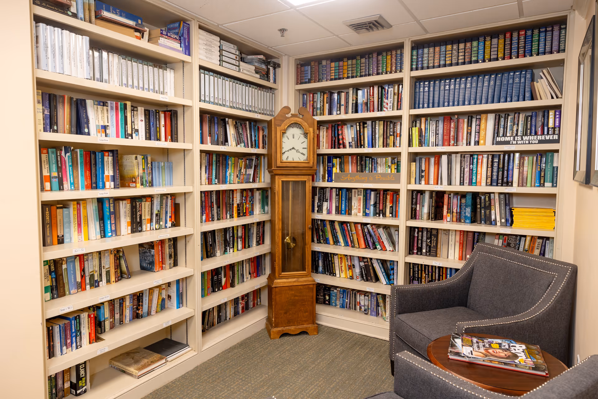 A cozy reading nook in a senior living facility featuring white bookshelves filled with books and binders, a wooden grandfather clock in the corner, and a gray upholstered armchair next to a small round wooden table with magazines on it.