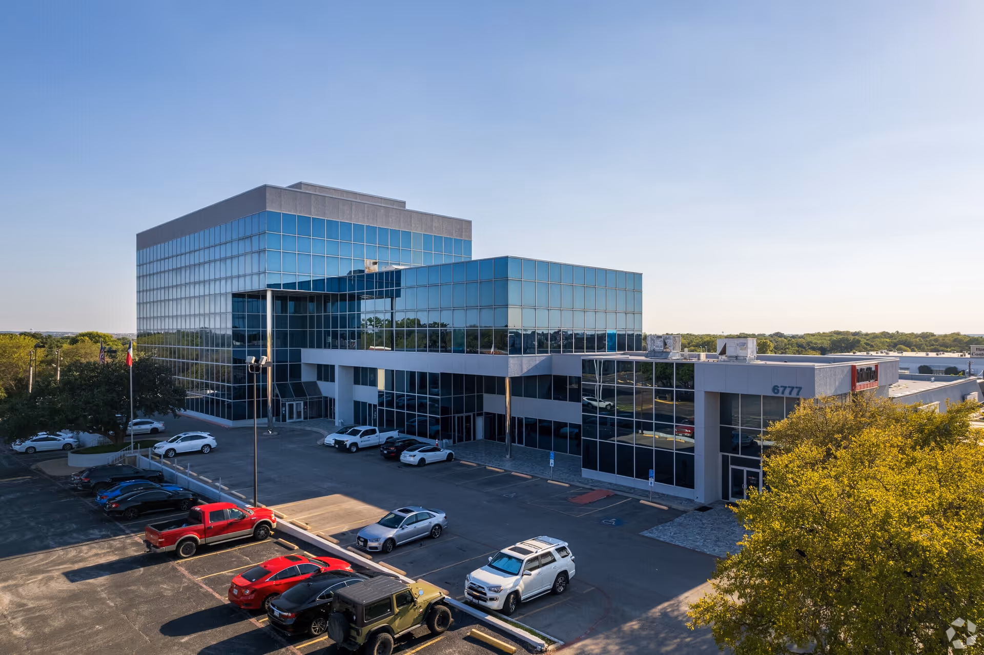 A modern glass-front office building with a parking lot and surrounding trees.