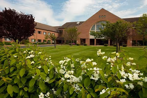 Front exterior of a brick senior living building with a manicured lawn, trees, and white flowering shrubs under a partly cloudy sky.