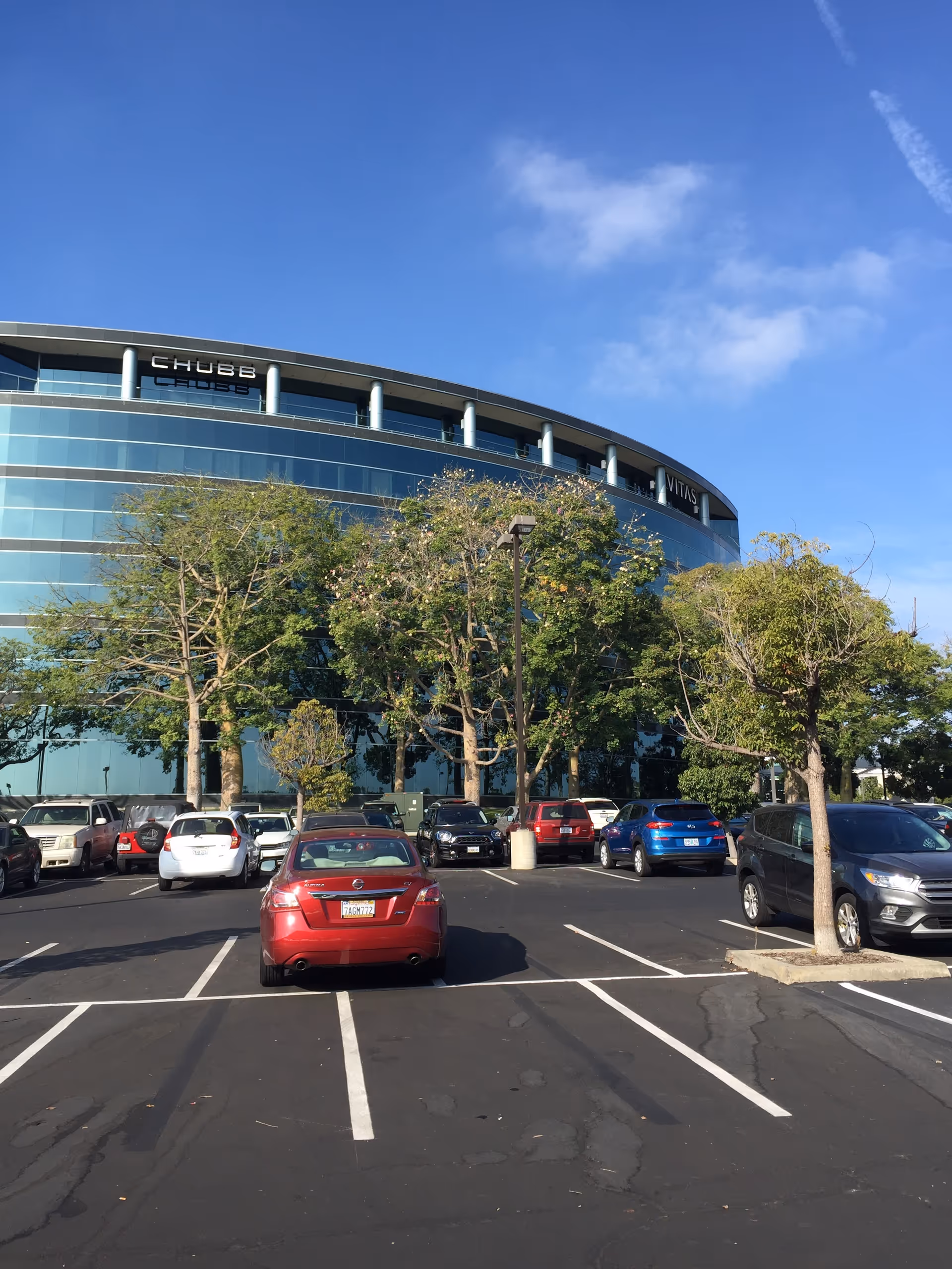 Curved glass exterior of a modern building with a parking lot and trees in foreground.