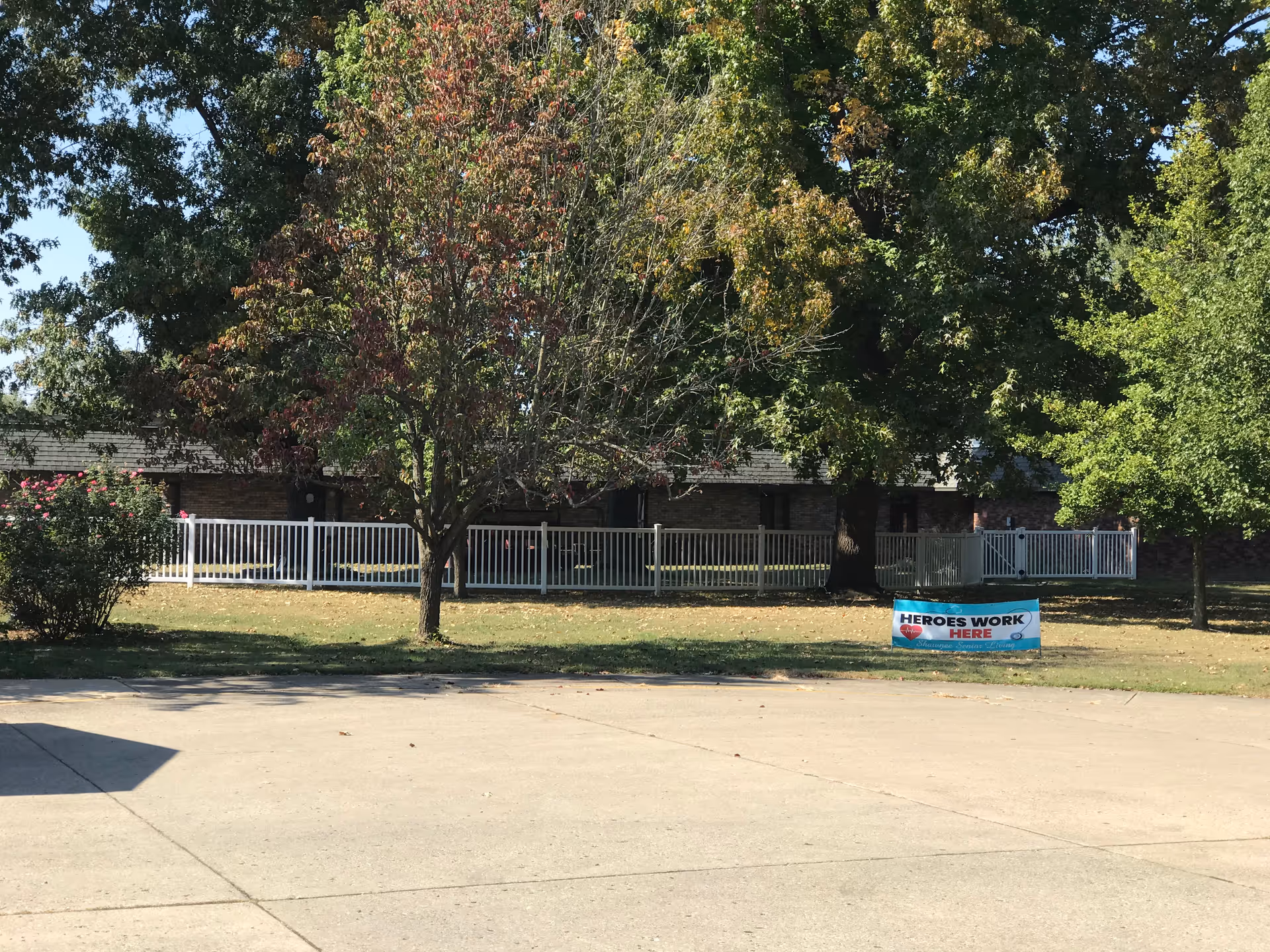 Outdoor view of a senior living facility with a paved area in the foreground, several trees with green and some autumn-colored leaves, a white fence, and a brick building in the background. A blue banner on the grass reads 'HEROES WORK HERE' with a red heart and additional smaller text.