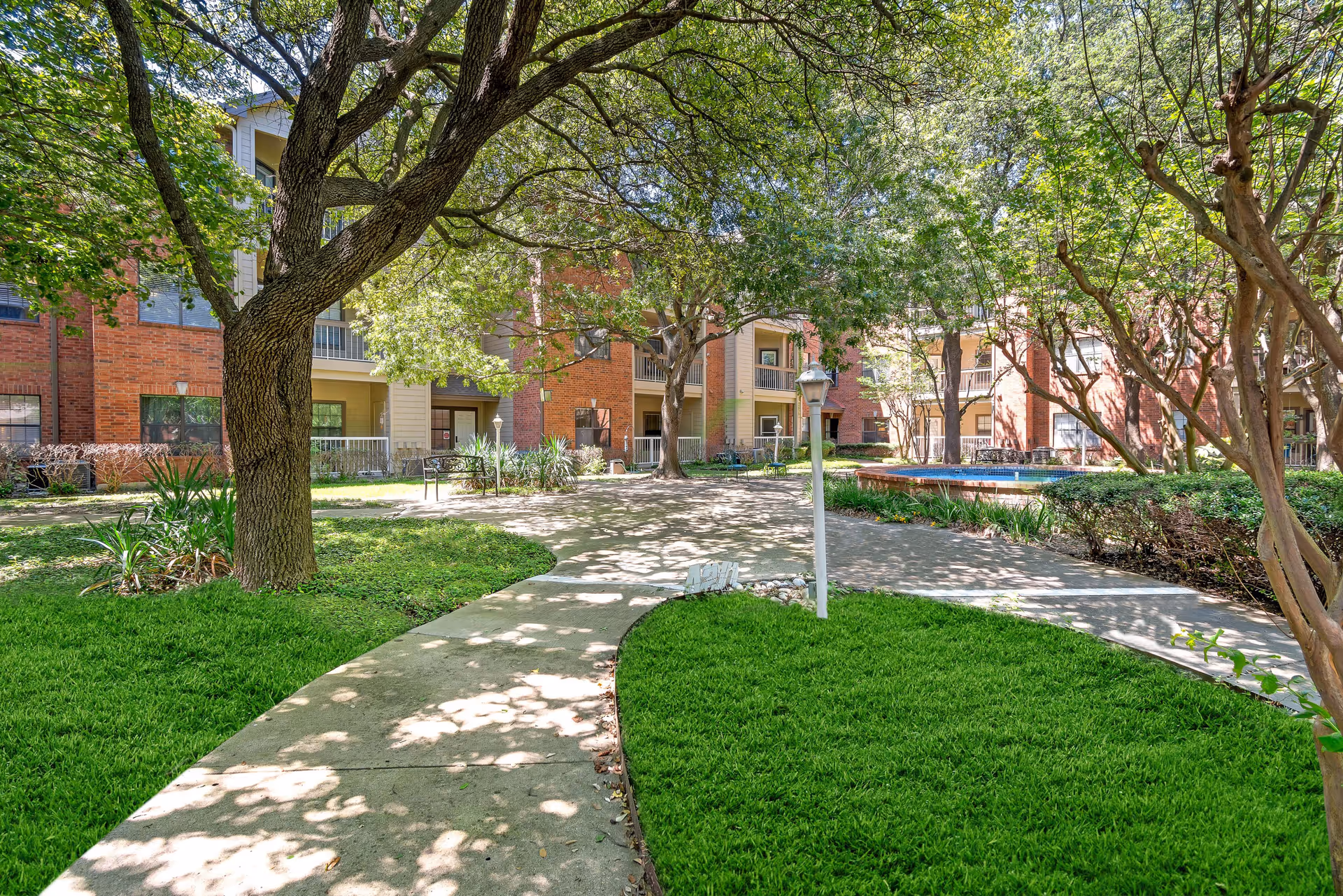 Outdoor courtyard area with a concrete walkway winding through green grass and trees, surrounded by a multi-story brick and beige building with balconies and windows. There are benches and a small fountain or water feature visible in the background.