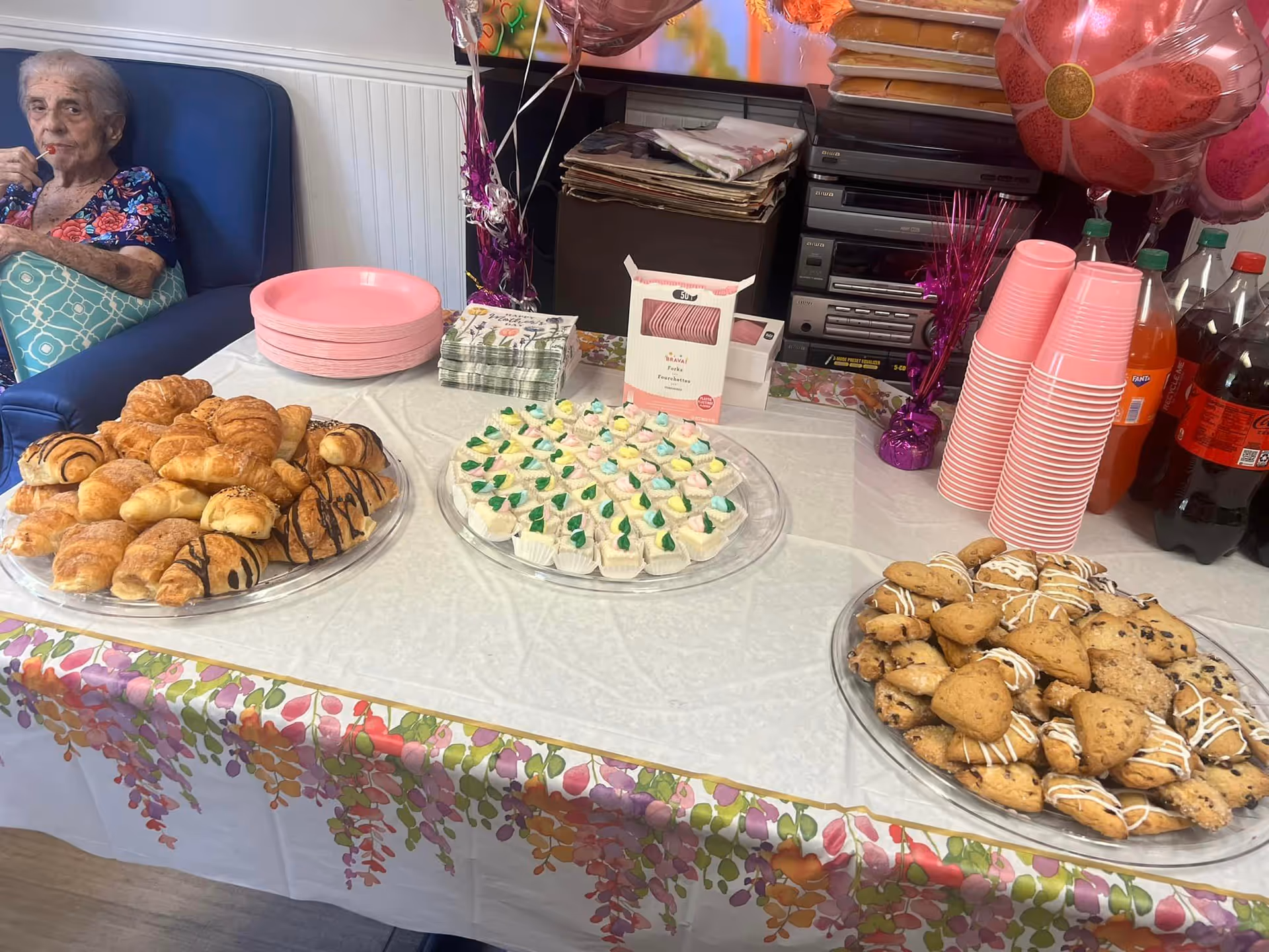 Elderly woman sits by a table of pastries at a celebration in an indoor dining area.