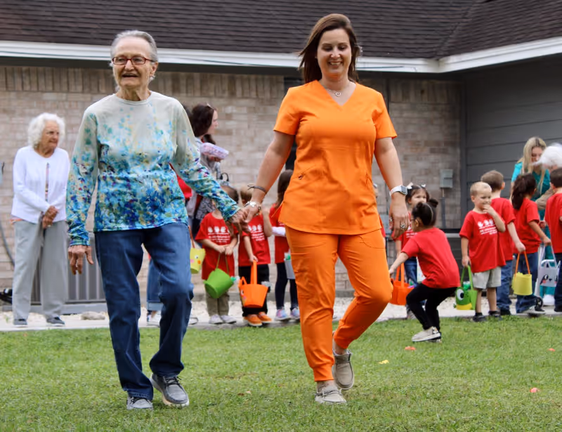 An elderly woman holding hands with a caregiver dressed in orange scrubs, walking on grass outside a building. In the background, a group of children wearing red shirts and holding colorful buckets are gathered near the building.