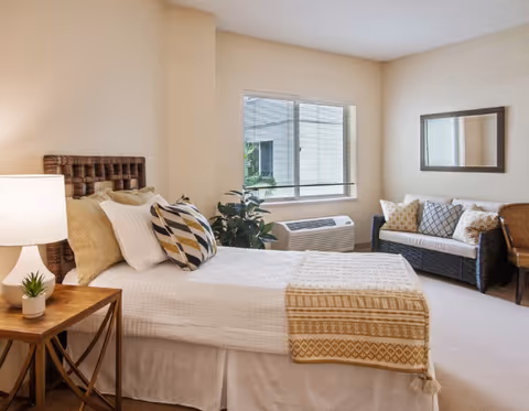 Bright bedroom in a senior living facility with a bed, sofa, window, and decor.