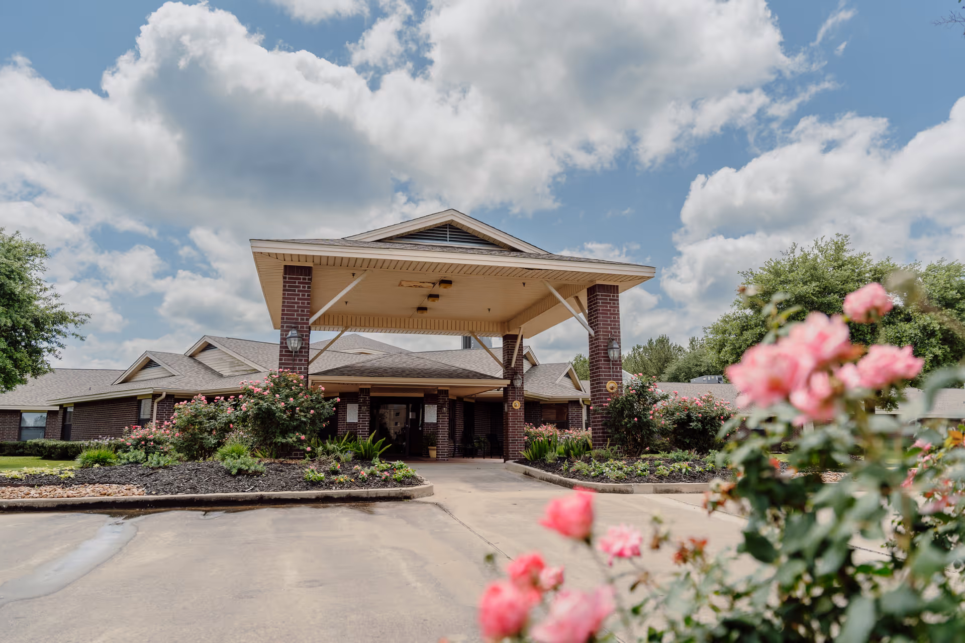 Front exterior view of Pine Ridge Health Care, LLP building with a covered entrance, brick pillars, landscaped flower beds, and pink flowers in the foreground under a partly cloudy sky.