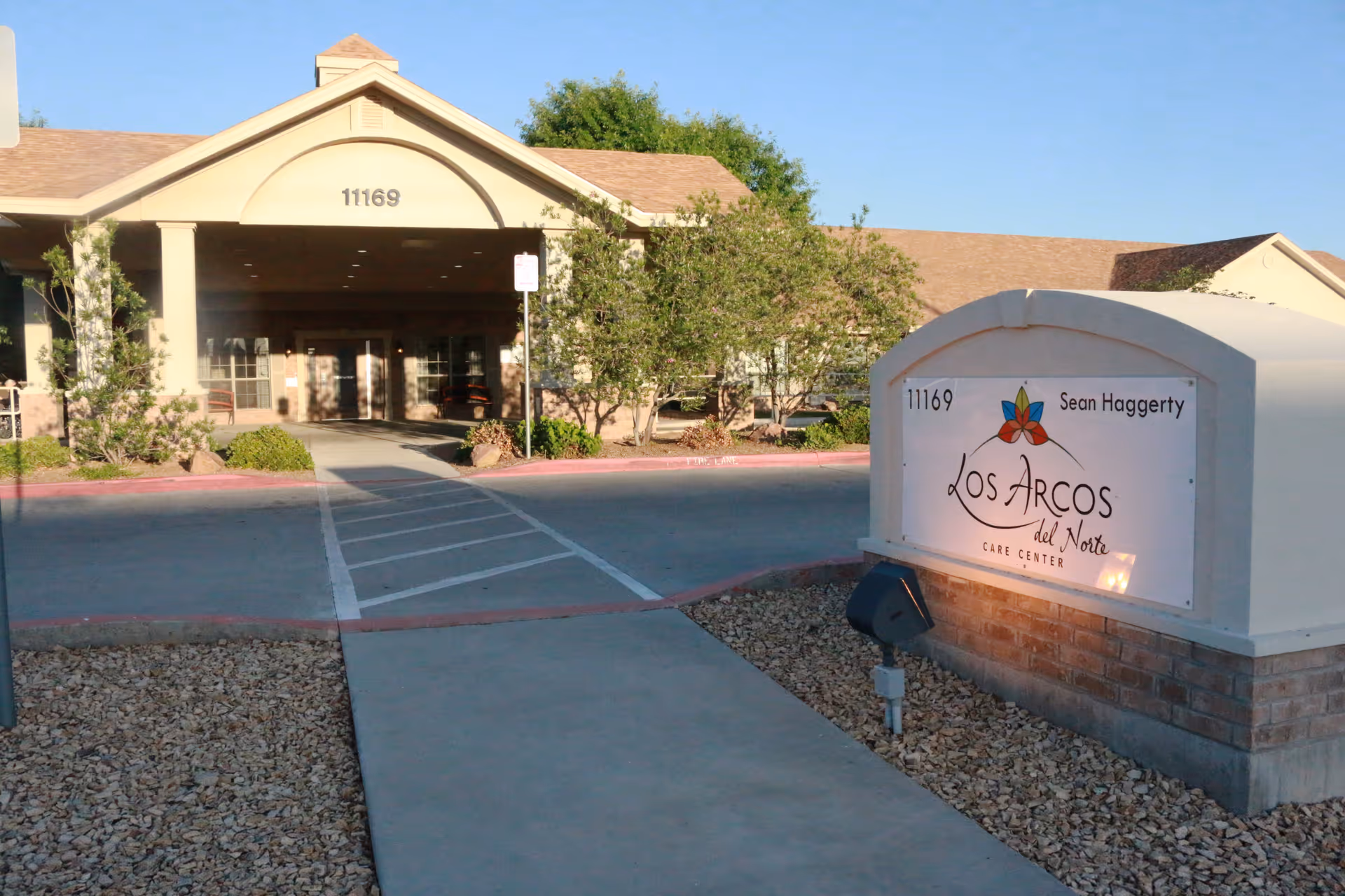 Exterior view of Los Arcos del Norte Care Center building with a sign displaying the facility name and address number 11169. The building has a covered entrance with columns, surrounded by trees and landscaping under a clear blue sky.